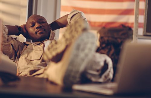Picture of a young african american soldier resting at his desk. His eyes are closed with his arms behind his head and his feet up on the desk. On the desk are a laptop and a small american flag. In the background is a bookshelf and on the wall a big american flag. Light is sipping threw a window from the left.